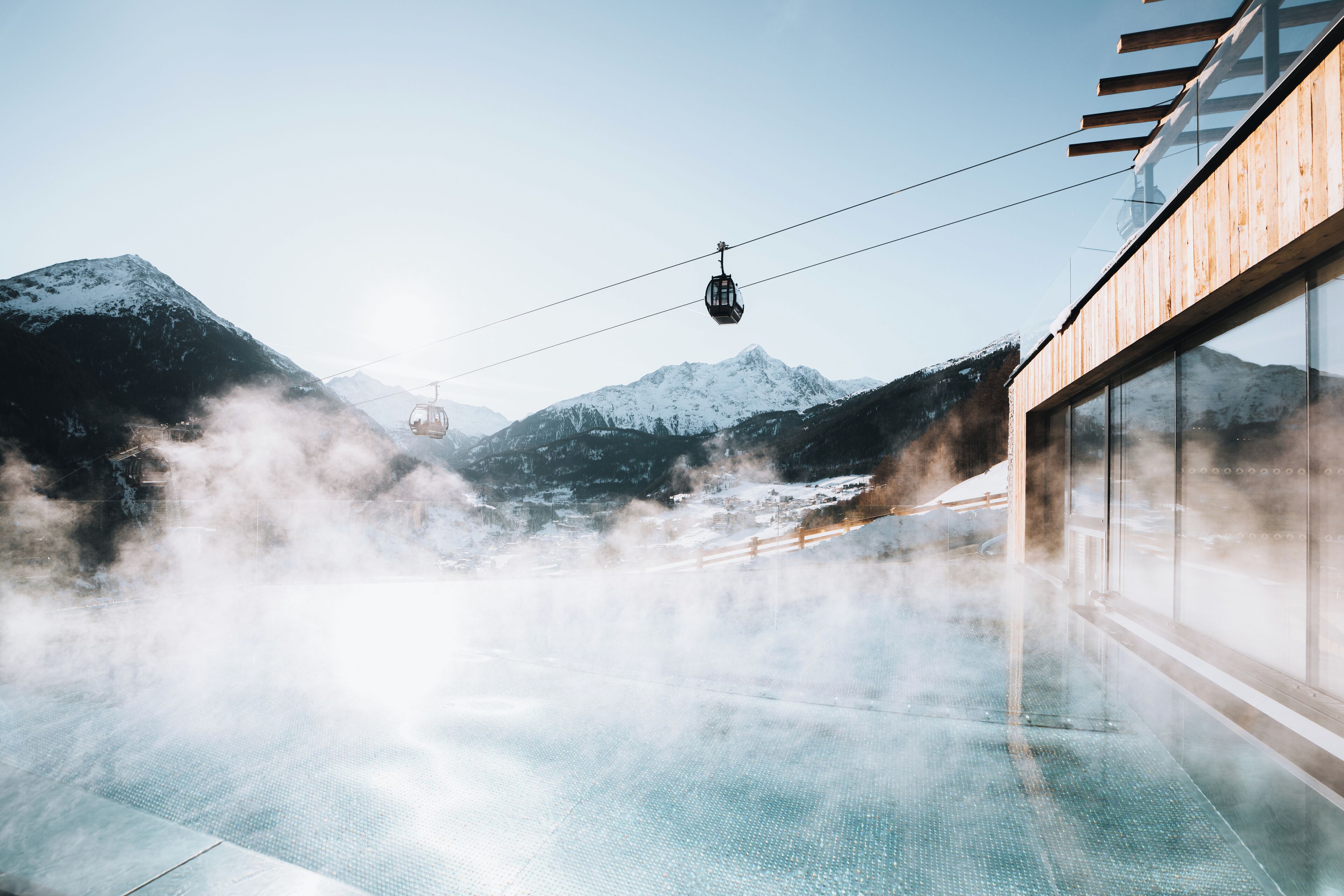 Un espace extérieur clos par des parois de verre, offrant une vue sur des montagnes enneigées, un téléphérique et la vapeur s'élevant du sol.
