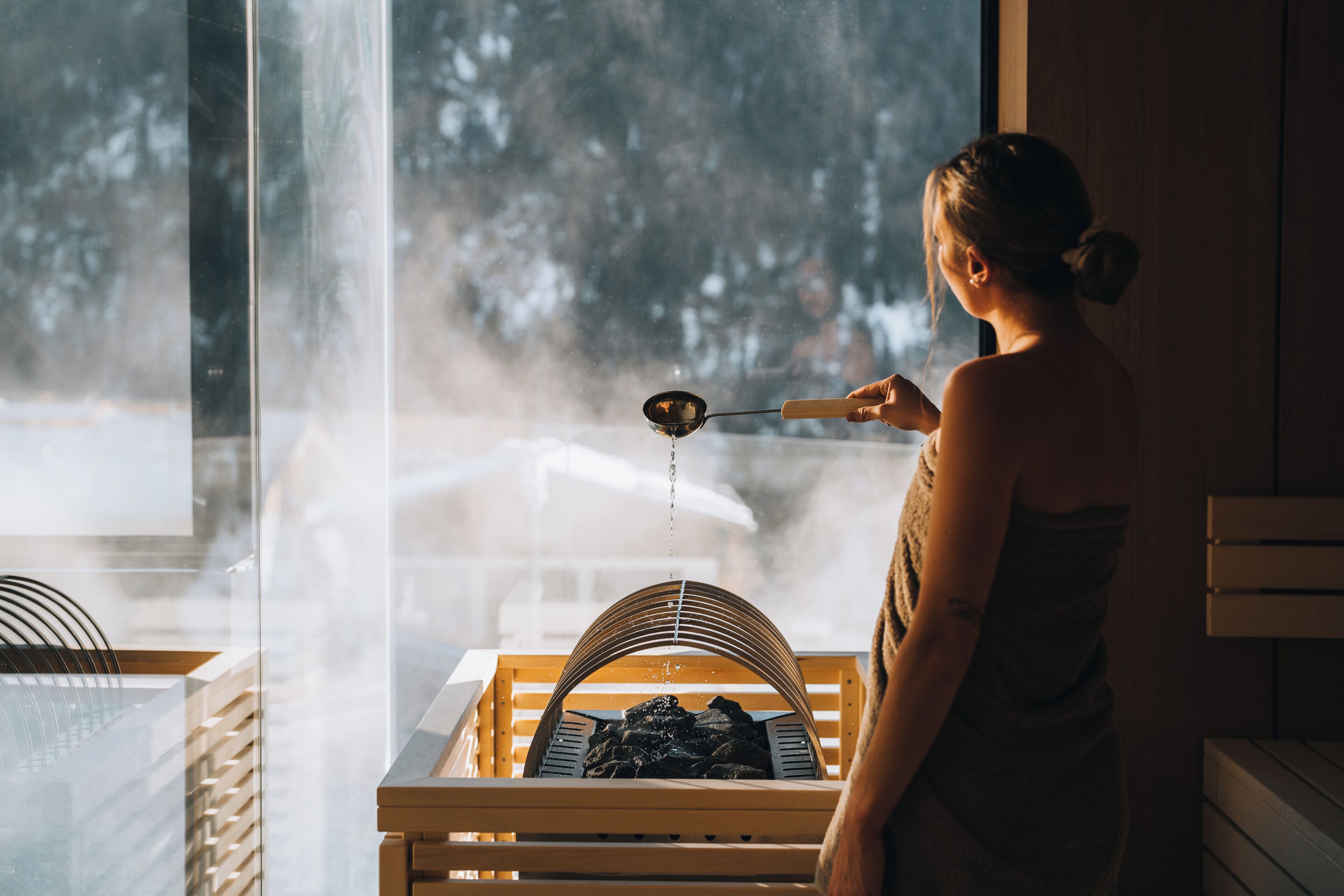 A person in a spa setting with a sauna, steam visible, and a window with a view of trees.