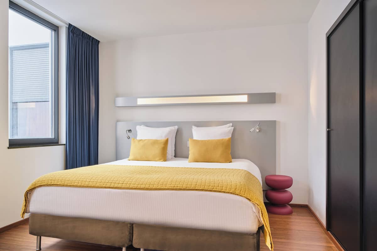 A modern hotel room with a large bed, a bedside table, a wardrobe, and a window with curtains.