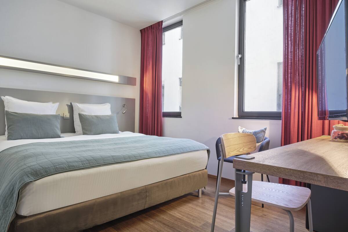A hotel room with a bed, a desk, and a window with curtains.