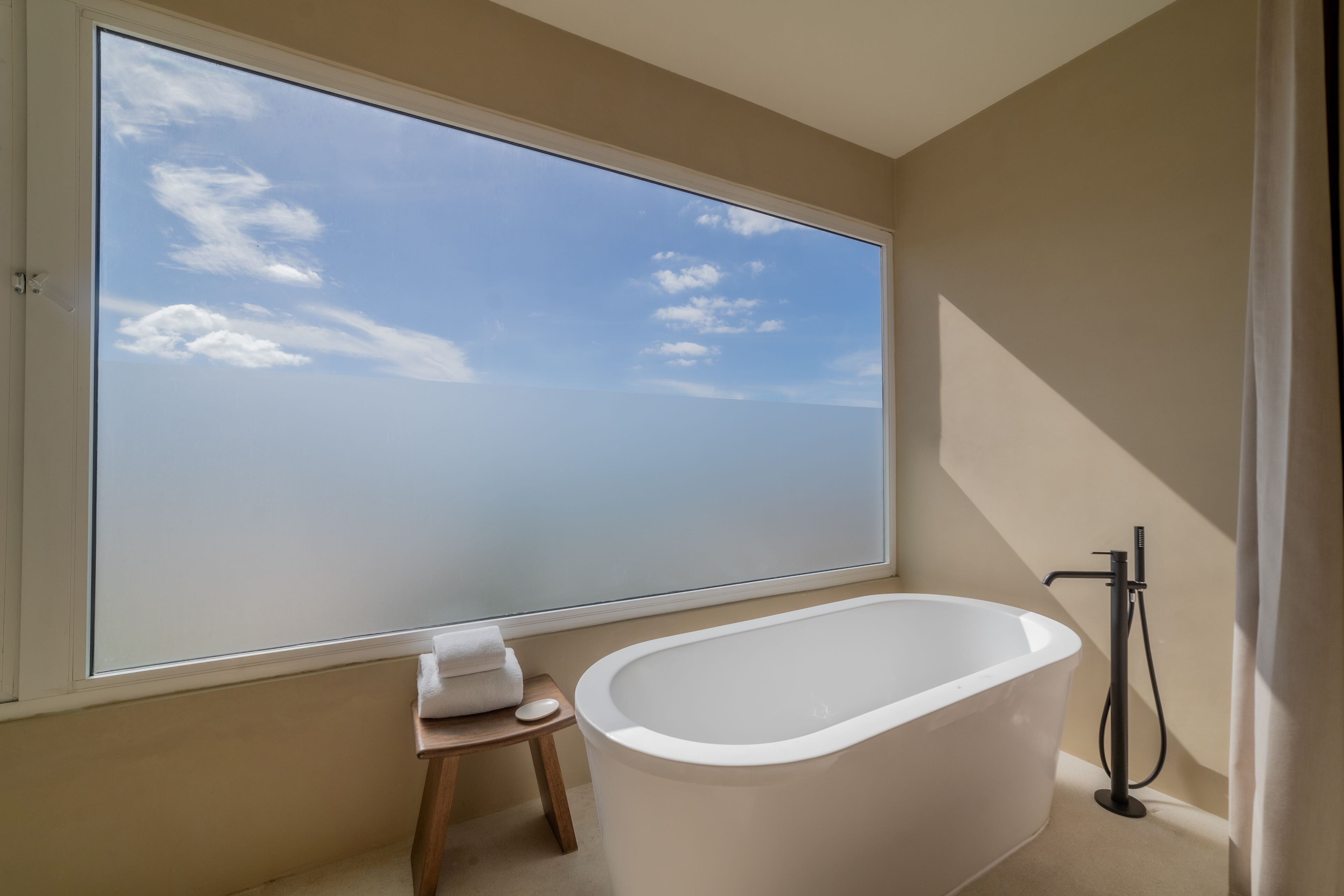 Un baño moderno con una bañera independiente, un taburete de madera pequeño y un ventanal con vistas al cielo.