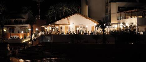 Un hotel frente al agua con fachada barroca, zona de piscina y barcos amarrados cerca.