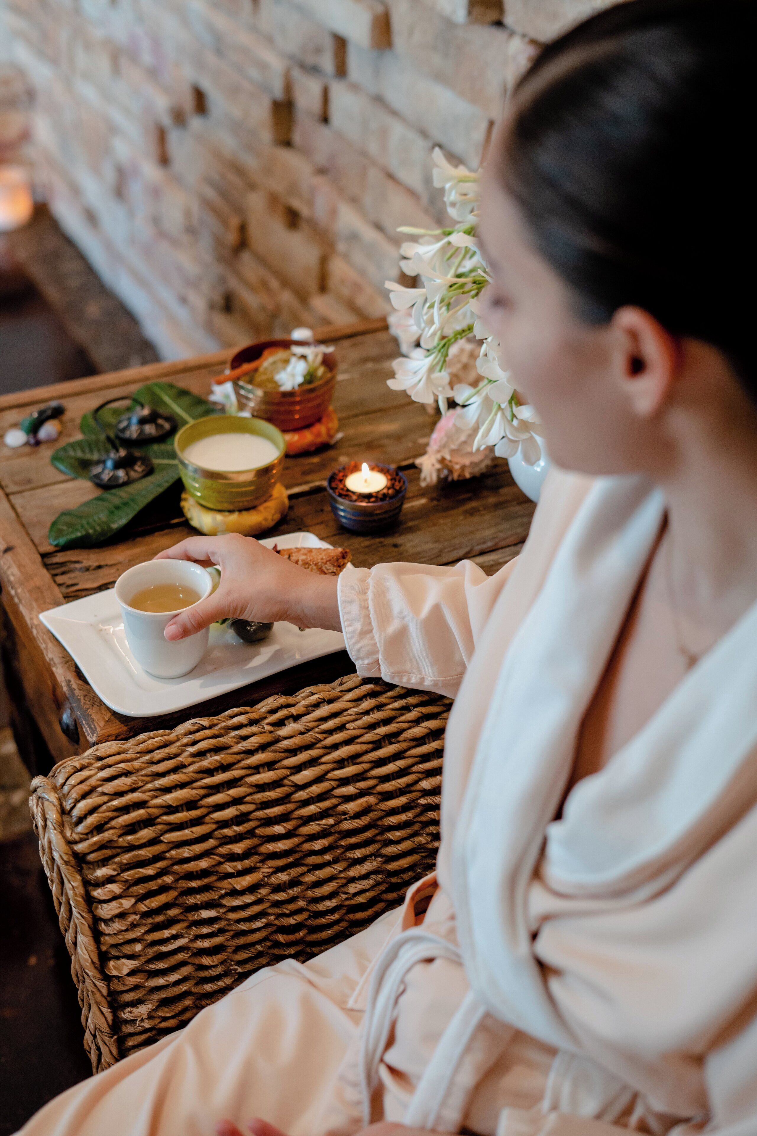 A person seated on a wicker chair at a wooden table with a cup of tea, a small bowl, and a candle.