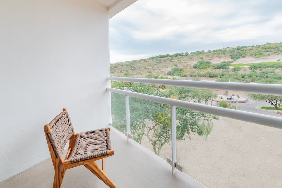 Un balcón con una silla de mimbre, barandilla blanca y vistas a un paisaje montañoso con vegetación escasa.