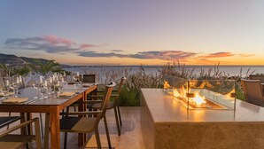 Un área de comedor al aire libre con vistas al mar al atardecer, que cuenta con una chimenea y mesas elegantemente dispuestas.