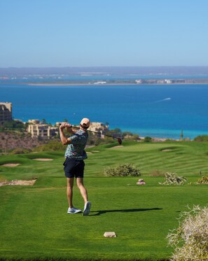 A person playing golf on a well-maintained course with a view of the ocean and buildings in the background.