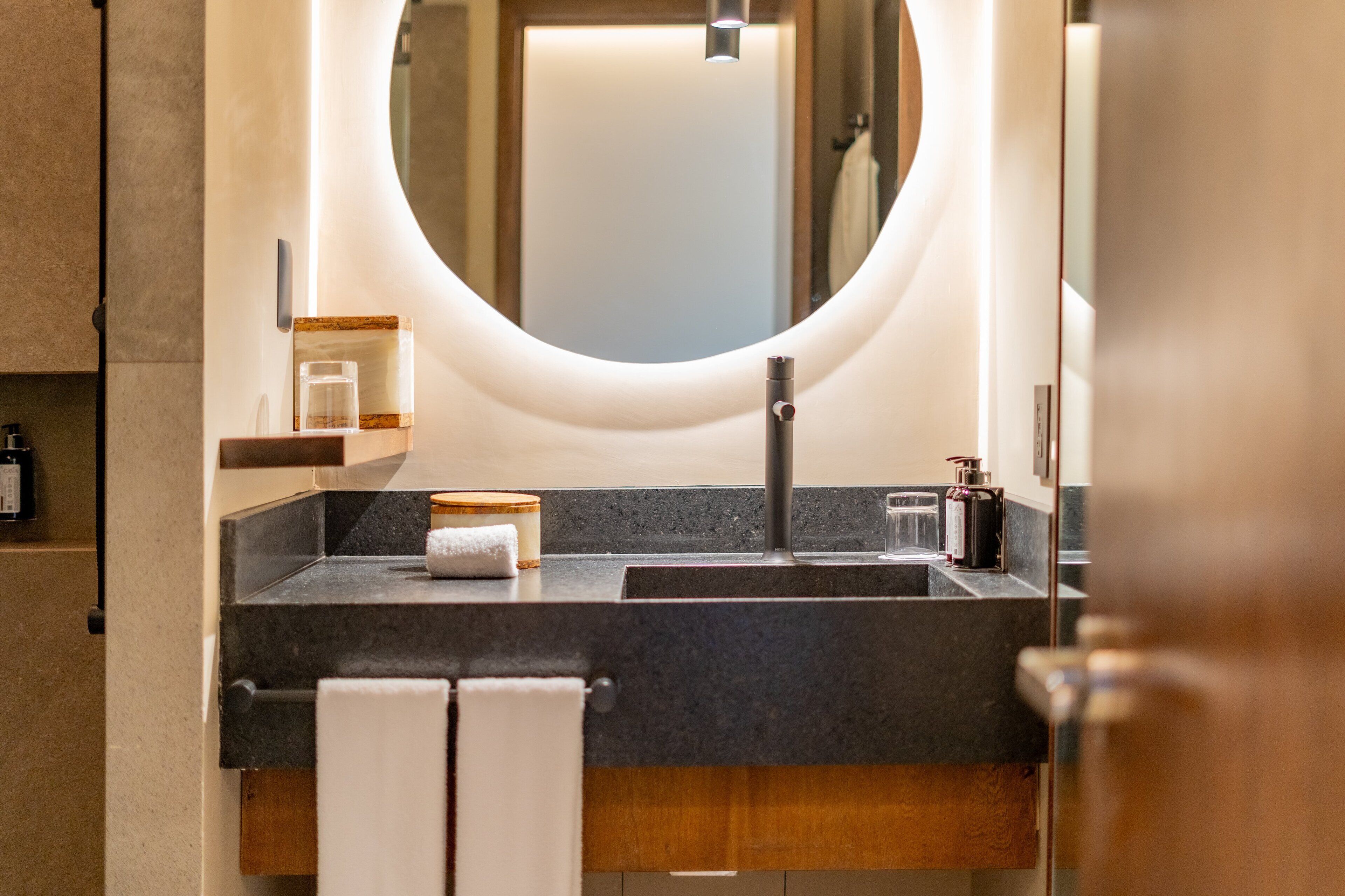 Une salle de bain moderne avec un grand miroir rond, deux vasques, un porte-serviettes et un distributeur de savon.