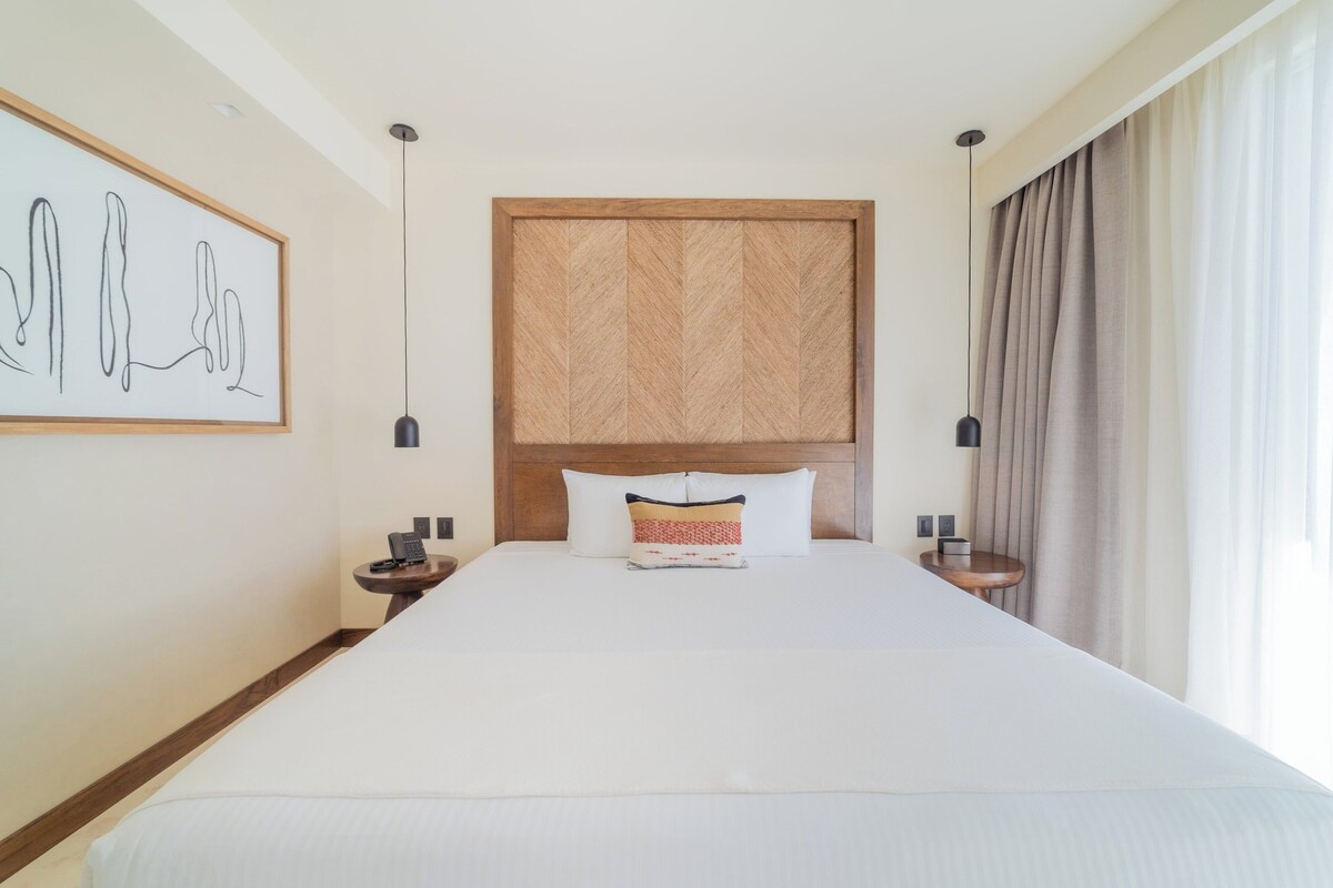 A large bed with a wooden headboard, two bedside tables with lamps, a framed artwork on the wall, and a window with curtains.