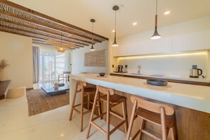 A modern kitchen with a central island, wooden bar stools, pendant lighting, and a large window with natural light.