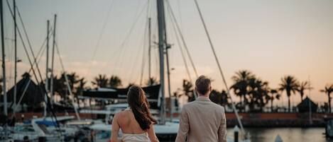 A couple holding hands at a marina with boats and palm trees in the background.