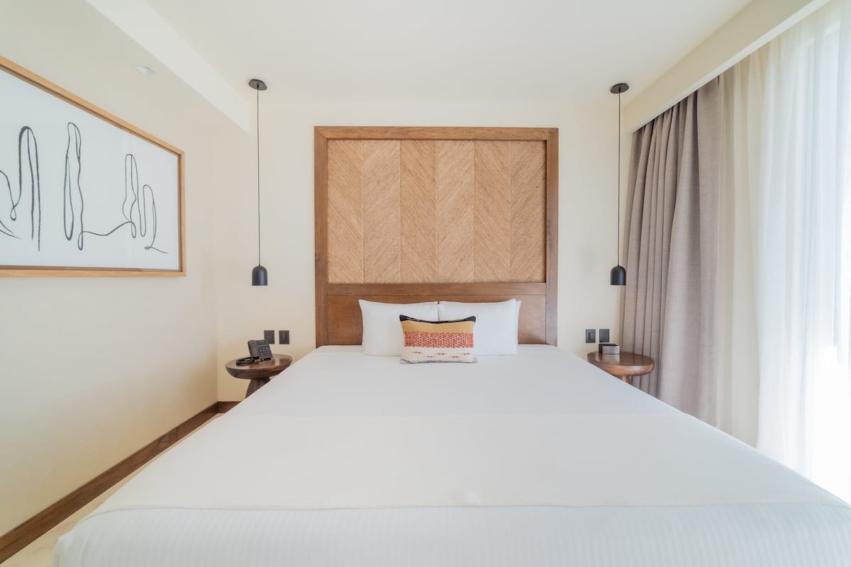 Una cama grande con cabecera de madera, dos mesitas de noche con lámparas, una obra de arte enmarcada en la pared y una ventana con cortinas.