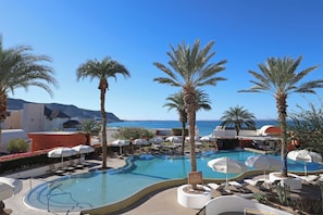 Zona de piscina en un resort con palmeras, sillones de descanso y sombrillas.