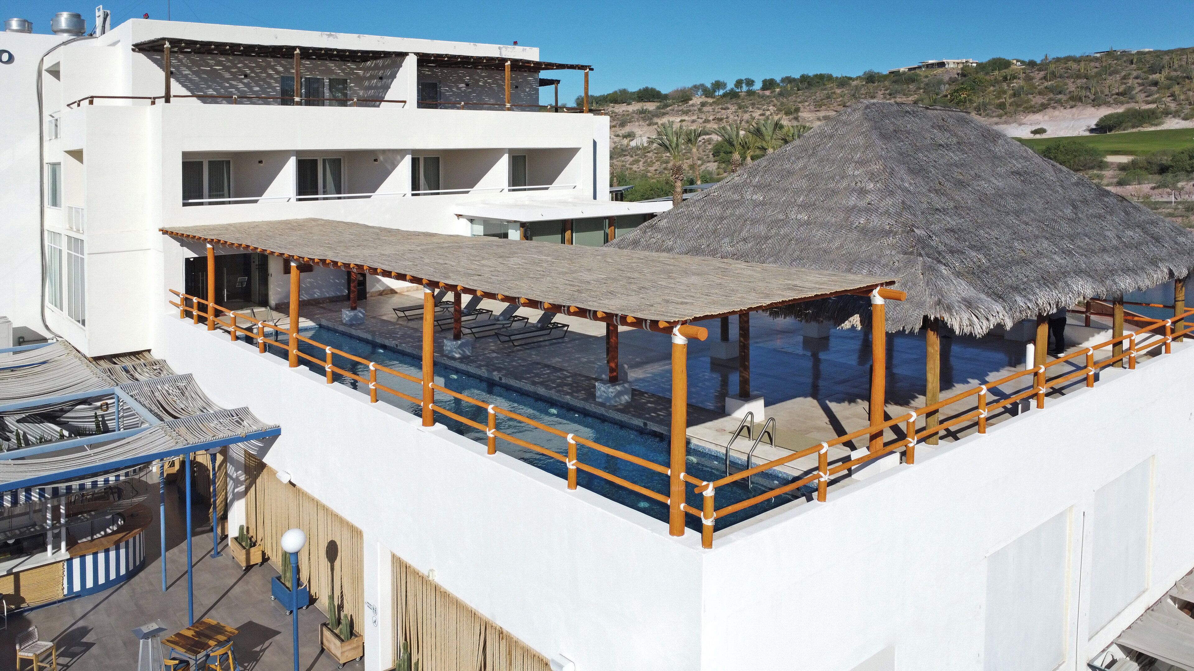 A rooftop pool with a thatched roof, a white building with multiple balconies, and a view of a hilly landscape.