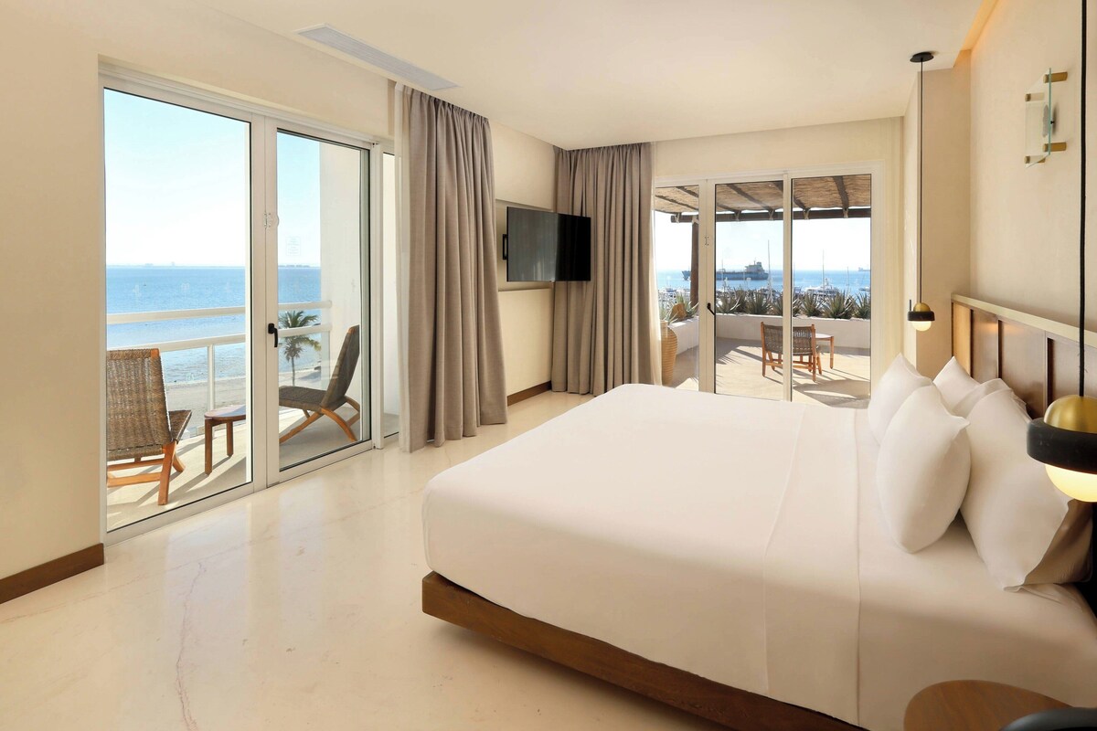 A hotel room with a large bed, a TV, a balcony with a view of the sea, and a dining area with chairs.