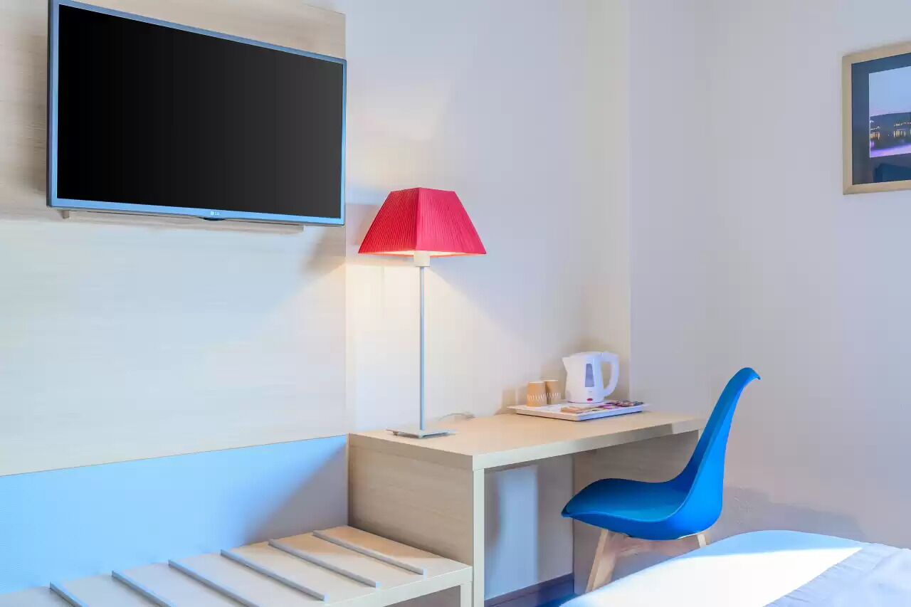Ein Hotelzimmer mit einem Bett, einem Schreibtisch mit Stuhl, einer roten Lampe, einem Flachbildfernseher und einem gerahmten Bild an der Wand.