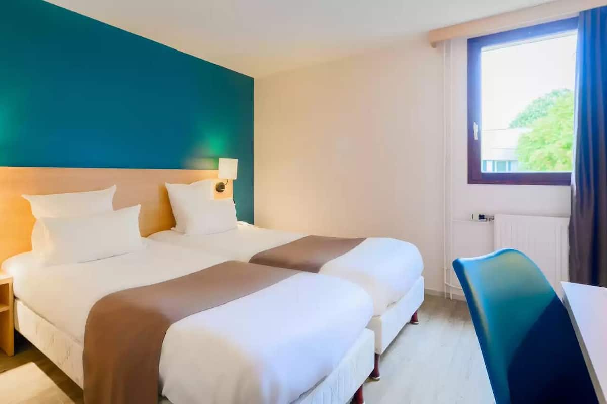 A hotel room with two beds, a desk, a chair, a window with curtains, and a teal accent wall.