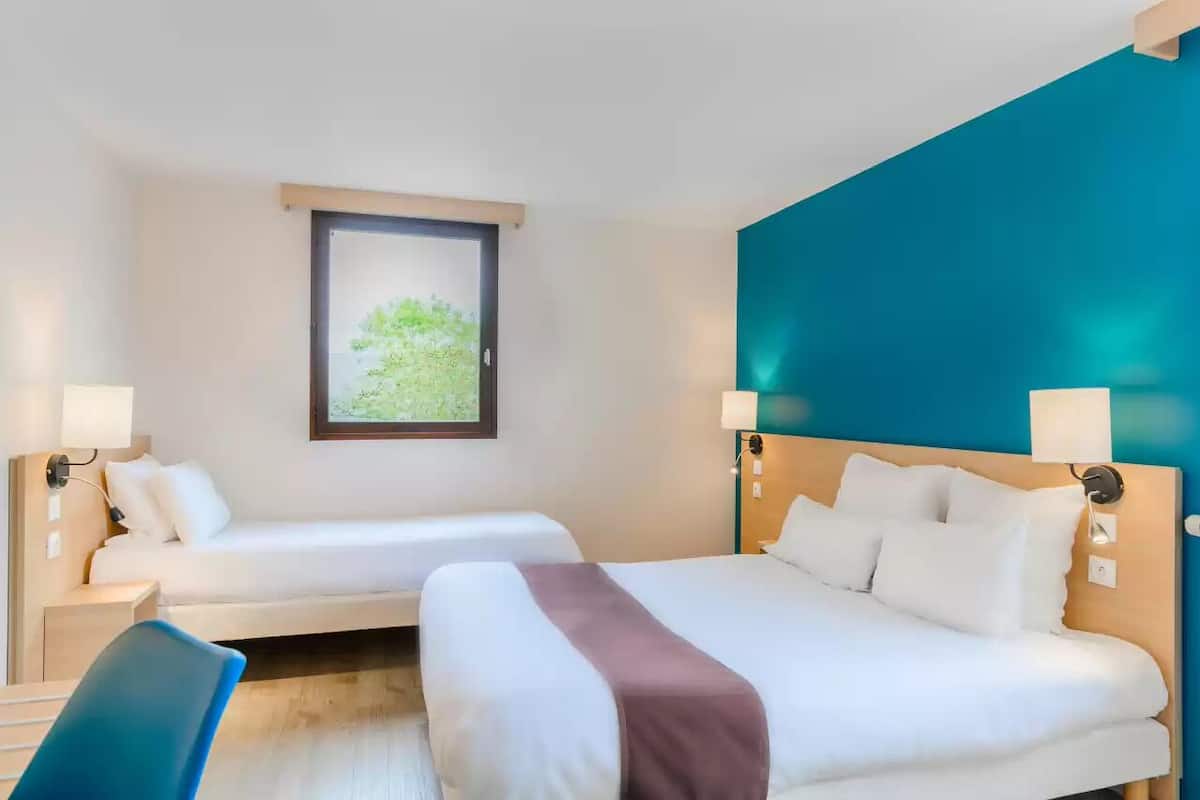 A hotel room with two beds, a desk, and a window with a view of greenery.