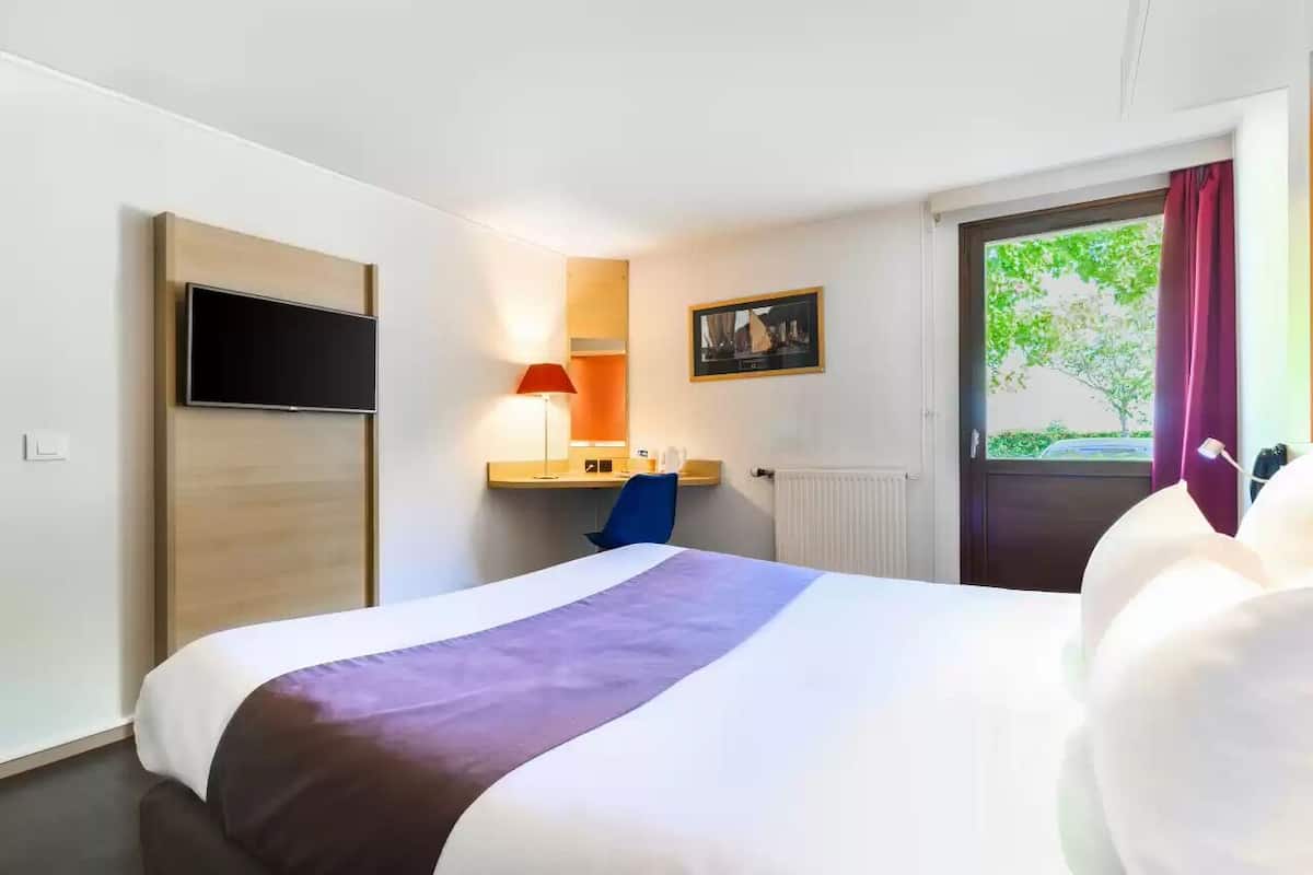 A hotel room with a bed, a desk with a chair, a TV mounted on the wall, a window with curtains, and a radiator.