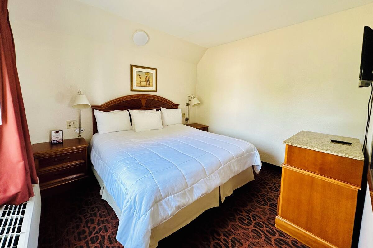 A hotel room with a large bed, two bedside tables, a wooden dresser, a ceiling light, and a framed picture on the wall.