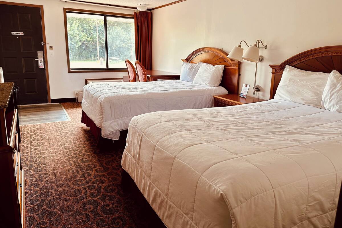 A hotel room with two beds, a desk, a chair, a window, and a door with a 'No Smoking' sign.