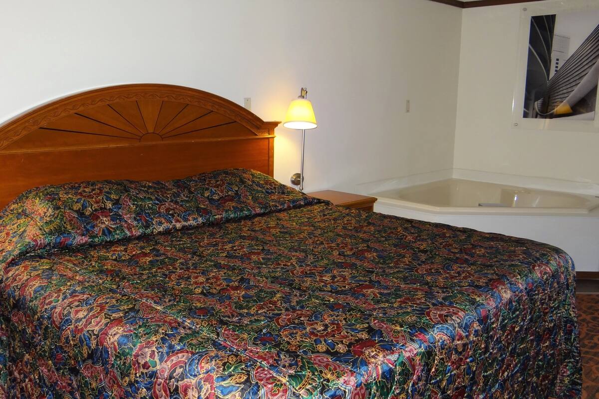 A hotel room with a wooden headboard, a colorful bedspread, a white bathtub, and two bedside lamps.