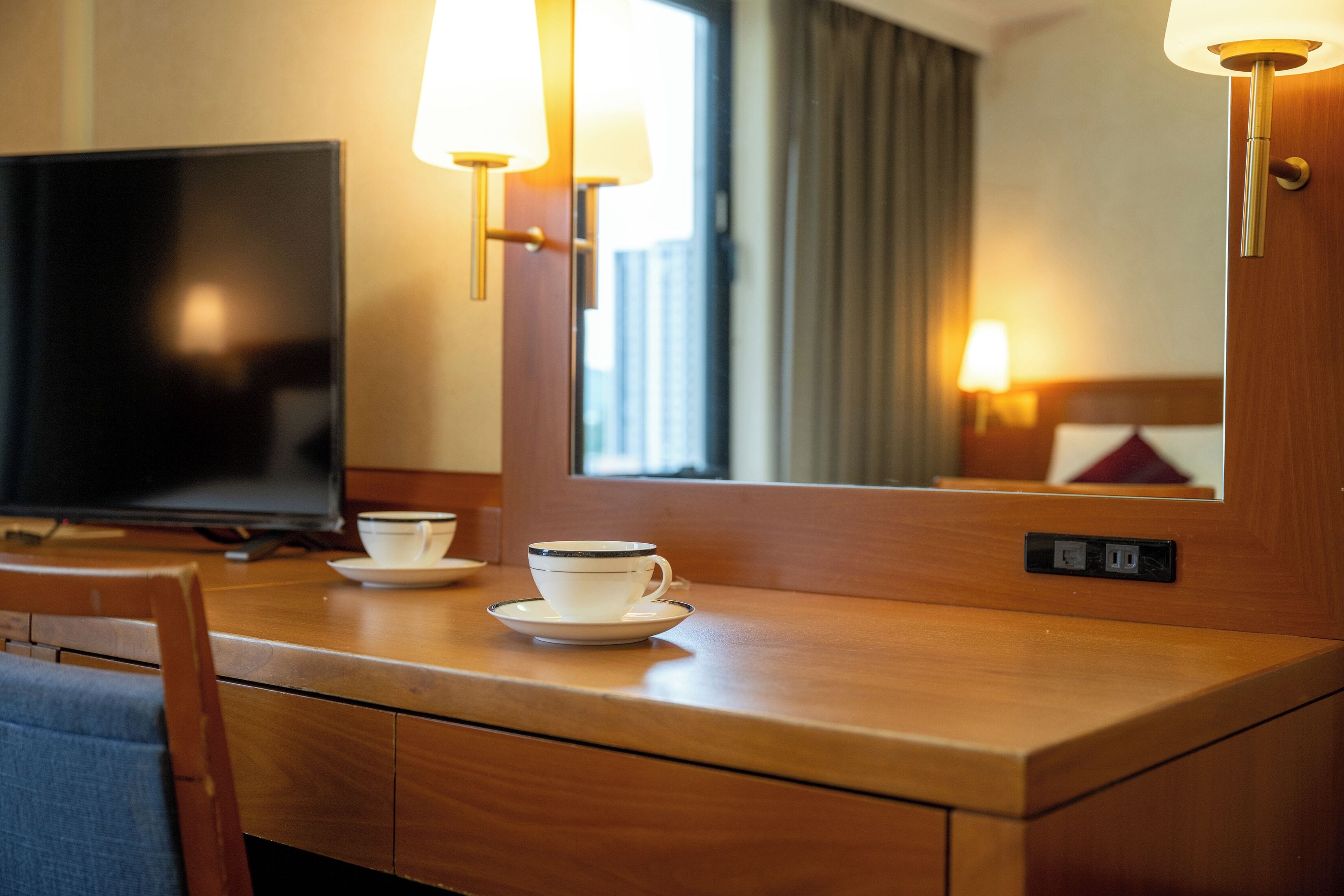 A hotel room with a wooden desk, a television, a cup and saucer, and a mirror reflecting a window view.