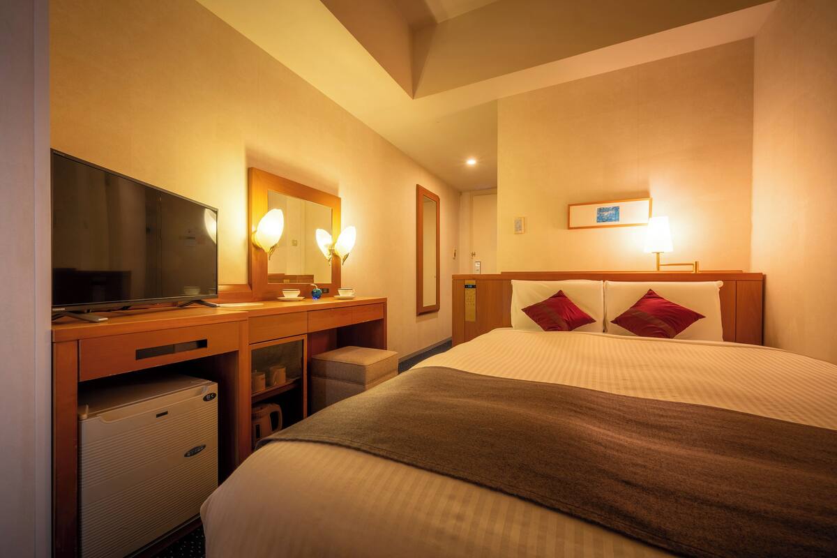 Una habitación de hotel con una cama, un escritorio con espejo, un televisor y un refrigerador pequeño.