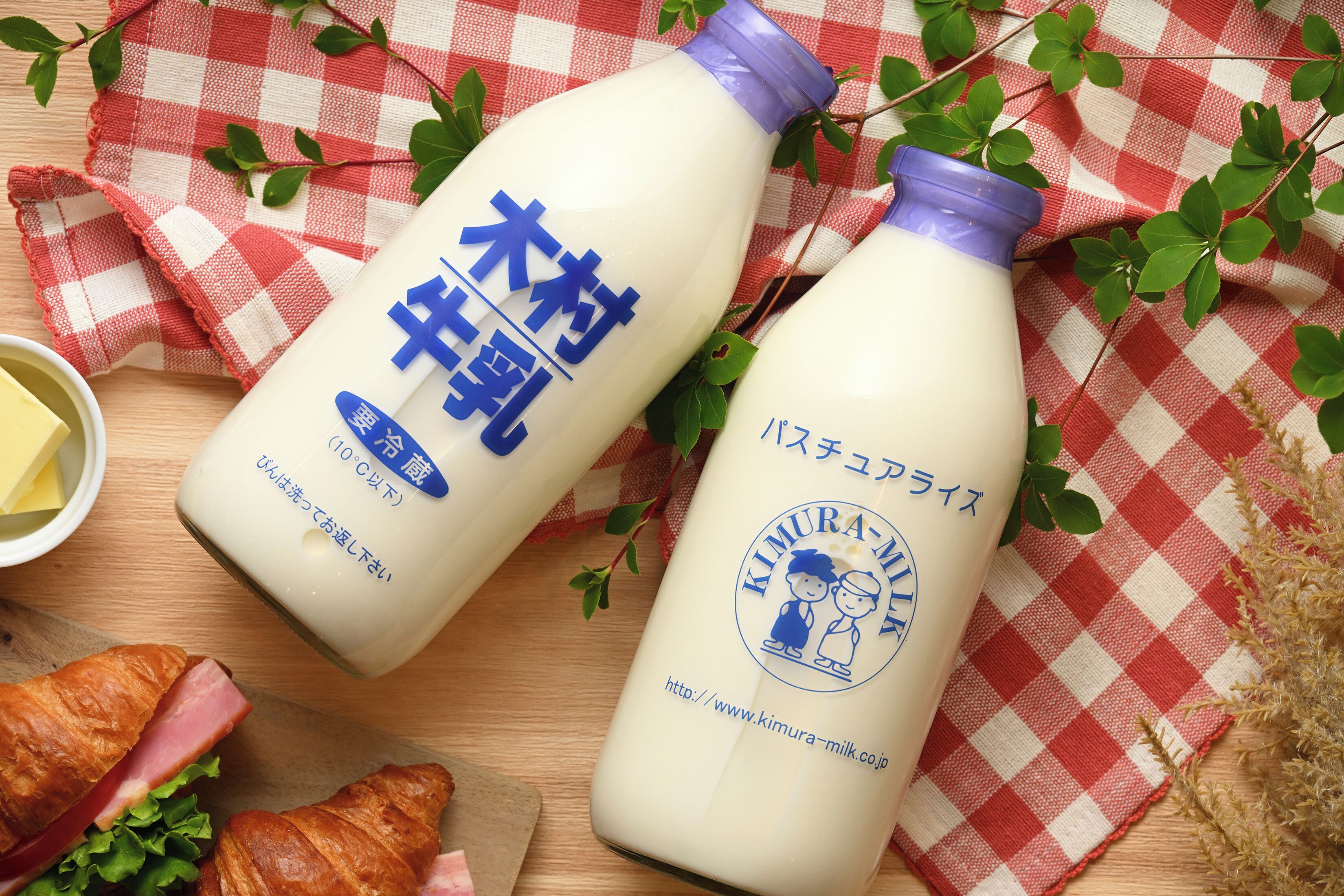 Two bottles of milk with labels, a croissant, and a small bowl of butter on a wooden surface with a red and white checkered cloth.