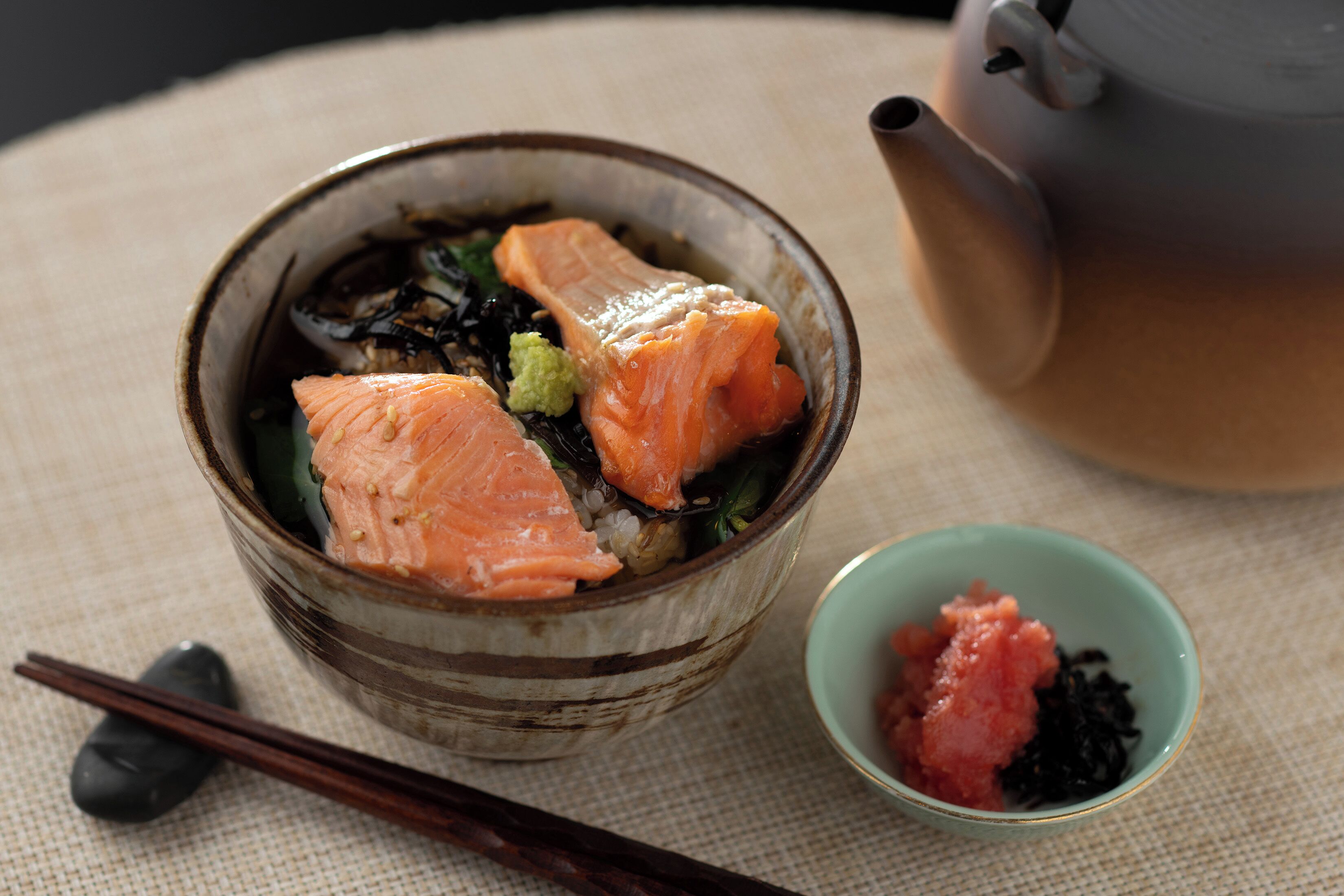 A bowl of salmon with seaweed and wasabi, a small bowl of wasabi, and chopsticks on a textured tablecloth.