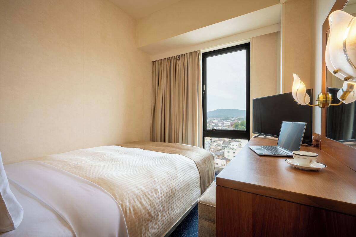 Una habitación de hotel con cama, un escritorio con una laptop y una taza, un ventanal con cortinas y vista a la ciudad.