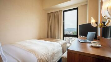A hotel room with a bed, a desk with a laptop and a cup, a large window with curtains, and a city view.