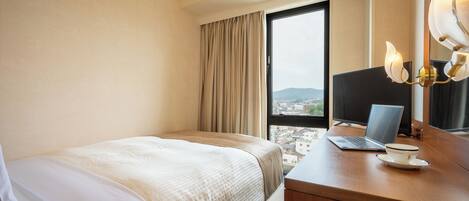 Una habitación de hotel con cama, un escritorio con una laptop y una taza, un ventanal con cortinas y vista a la ciudad.