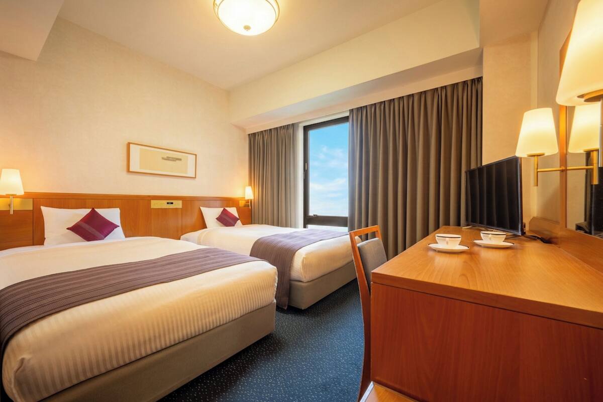 Una habitación de hotel con dos camas, un escritorio, una silla, un televisor y una ventana con cortinas.