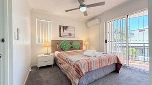 A bedroom with a bed, two bedside lamps, a ceiling fan, a window with blinds, a painting on the wall, and a view of the outside through the window. - Santana Resort (Surfers Paradise)