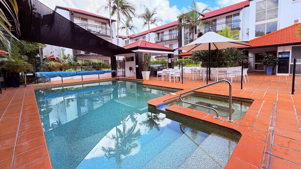 A swimming pool with a tiled deck, surrounded by lounge chairs and umbrellas, with a building in the background. - Santana Resort (Surfers Paradise)