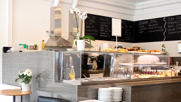 A modern cafe with a counter displaying food items, a seating area with a table and chairs, and a chalkboard menu. - Hotel Alexander (Maenttae-Vilppula)