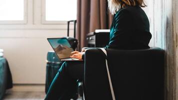 A person sitting on a chair in a room with a laptop, a small black bag, and a window with curtains.
