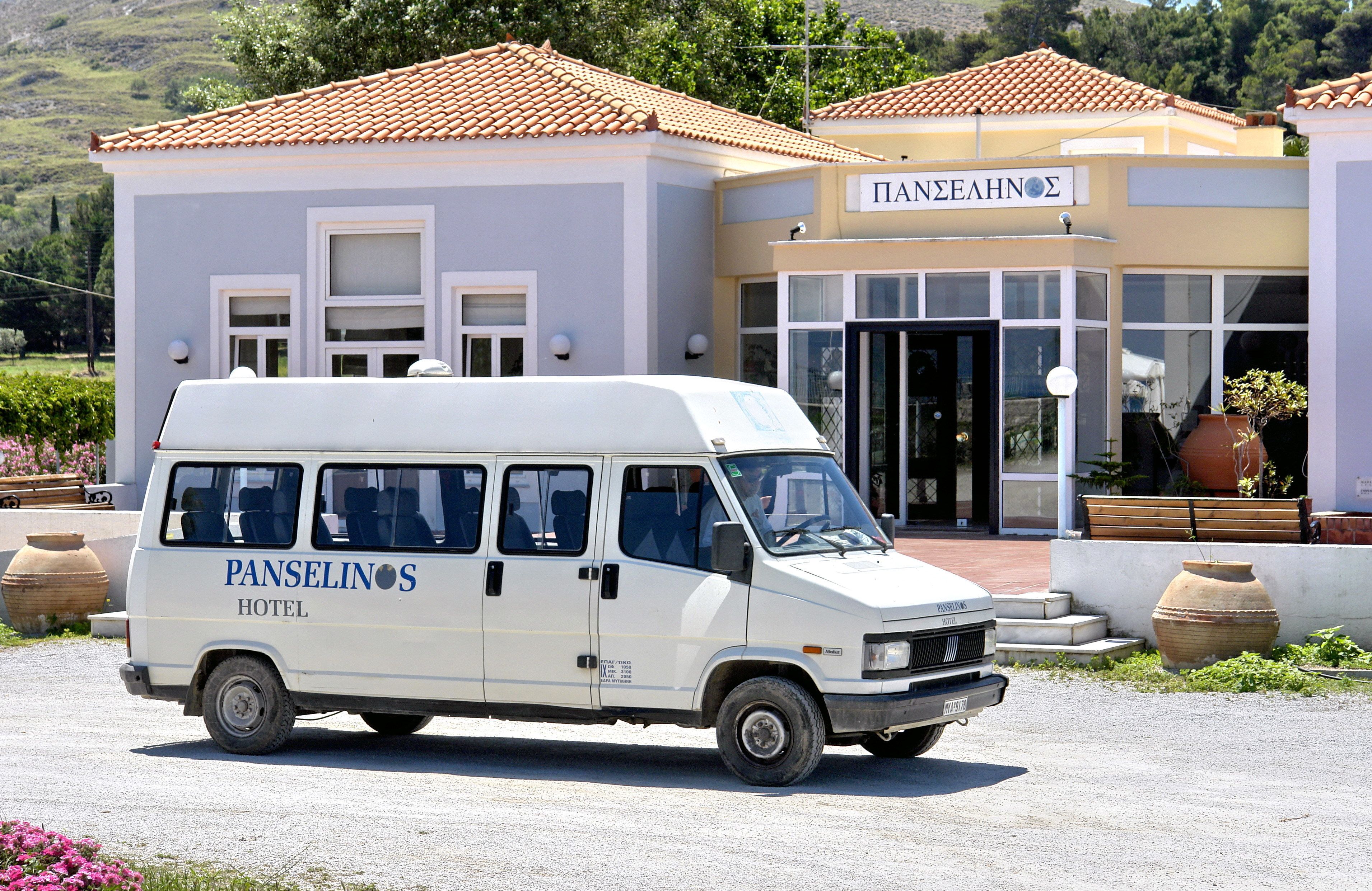 A white van with "Panselinos Hotel" written on the side parked in front of a building with a tiled roof and a sign that reads "PANEAHNOZ".
