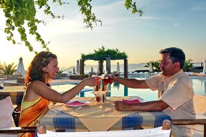 Una pareja brindando en una mesa junto a una piscina, con vista a la piscina y montañas al fondo.