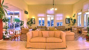 A spacious living area with multiple seating options, large windows offering views, and indoor plants. - Panselinos Hotel (Lesvos)