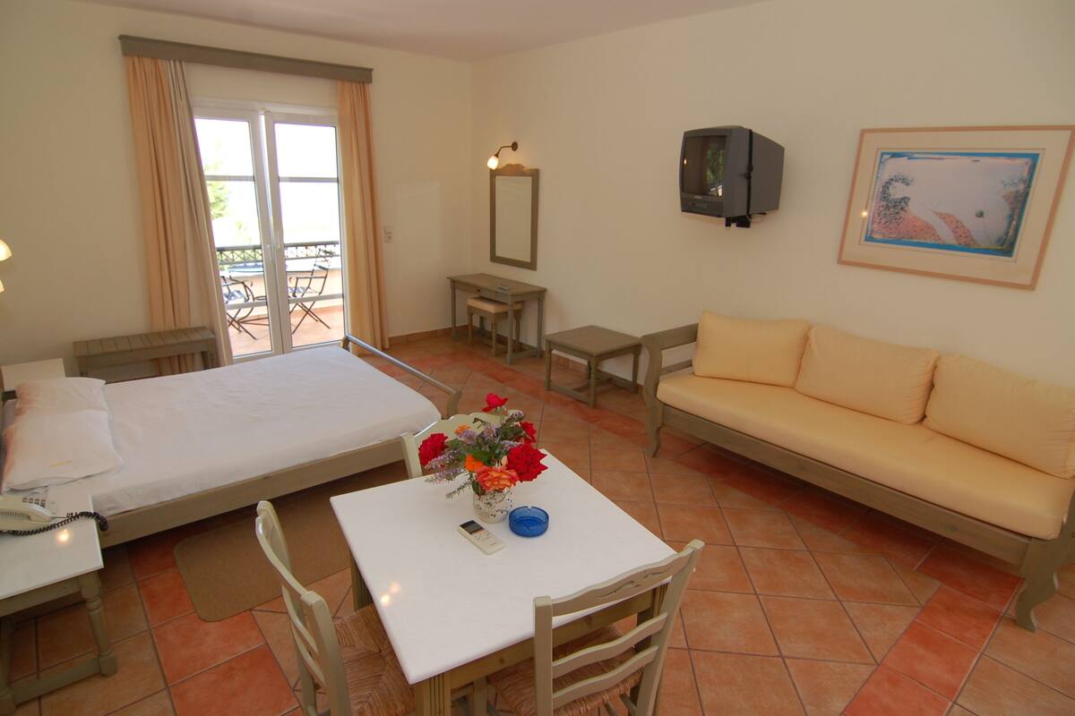 A hotel room with a bed, a sofa, a table with a vase of flowers, a desk with a lamp, a chair, a TV, and a balcony with chairs.
