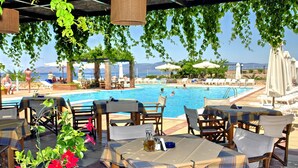 An outdoor pool area with lounge chairs and tables, surrounded by greenery and overlooking the sea. - Panselinos Hotel (Lesvos)