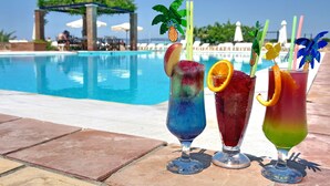 Three colorful cocktails with fruit garnishes on a poolside table, with a pool and lounge area in the background. - Panselinos Hotel (Lesvos)