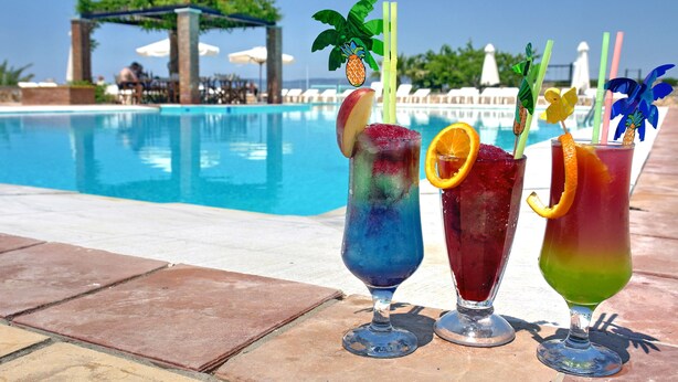 Three colorful cocktails with fruit garnishes on a poolside table, with a pool and lounge area in the background. - Panselinos Hotel (Lesvos)