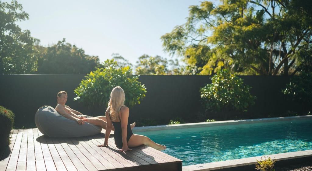 A couple sitting on a wooden deck by a swimming pool with trees in the background.