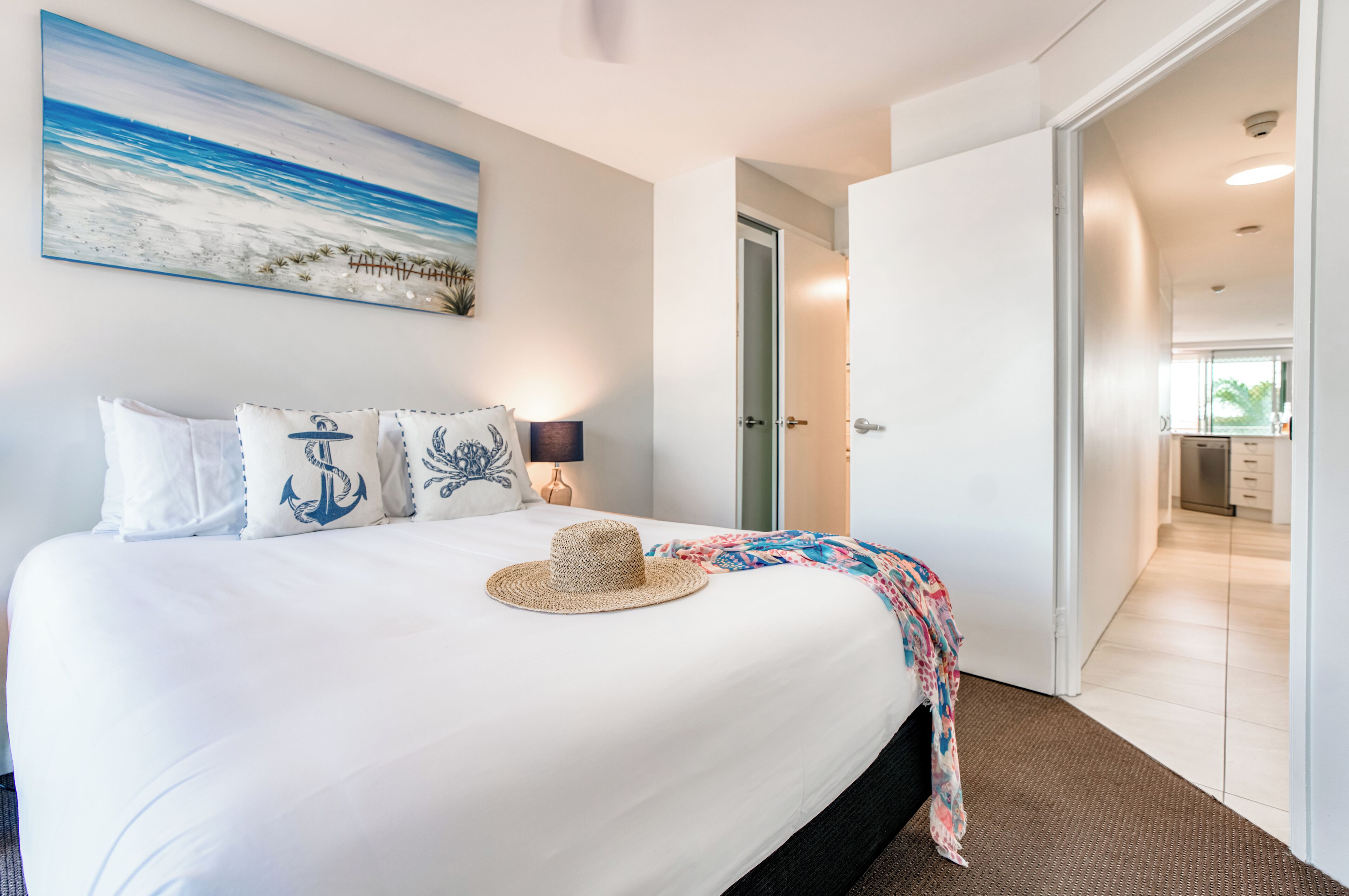 Ein ordentlich bezogenes Bett mit einem Strohhut, Kissen mit Anker-Motiven und einem Strandbild an der Wand.