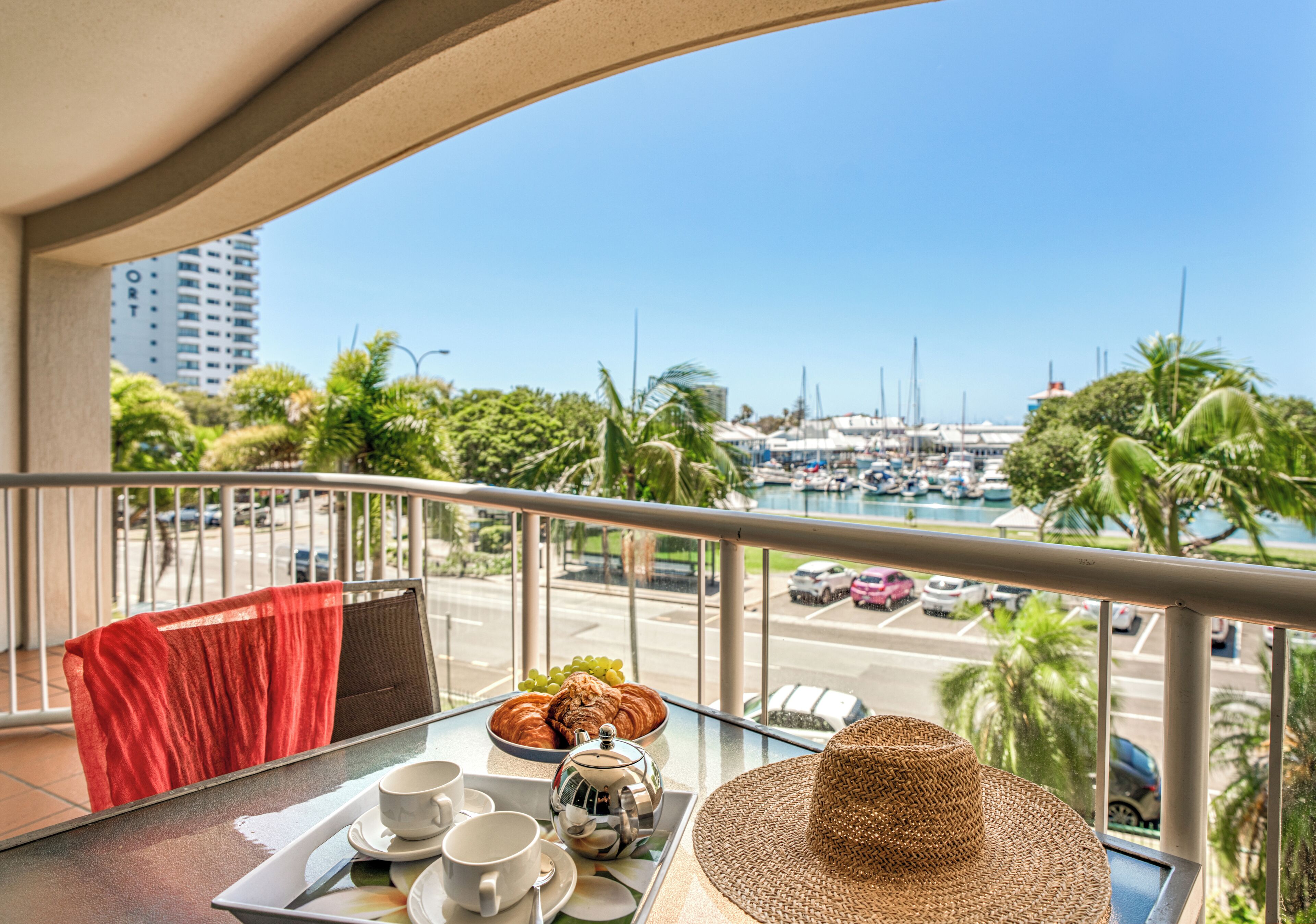 A balcony with a table set for breakfast, overlooking a marina with boats and palm trees.