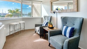 A modern living room with a large window, two bar stools, a grey armchair, a floor lamp, and a small table with a bowl of fruit.