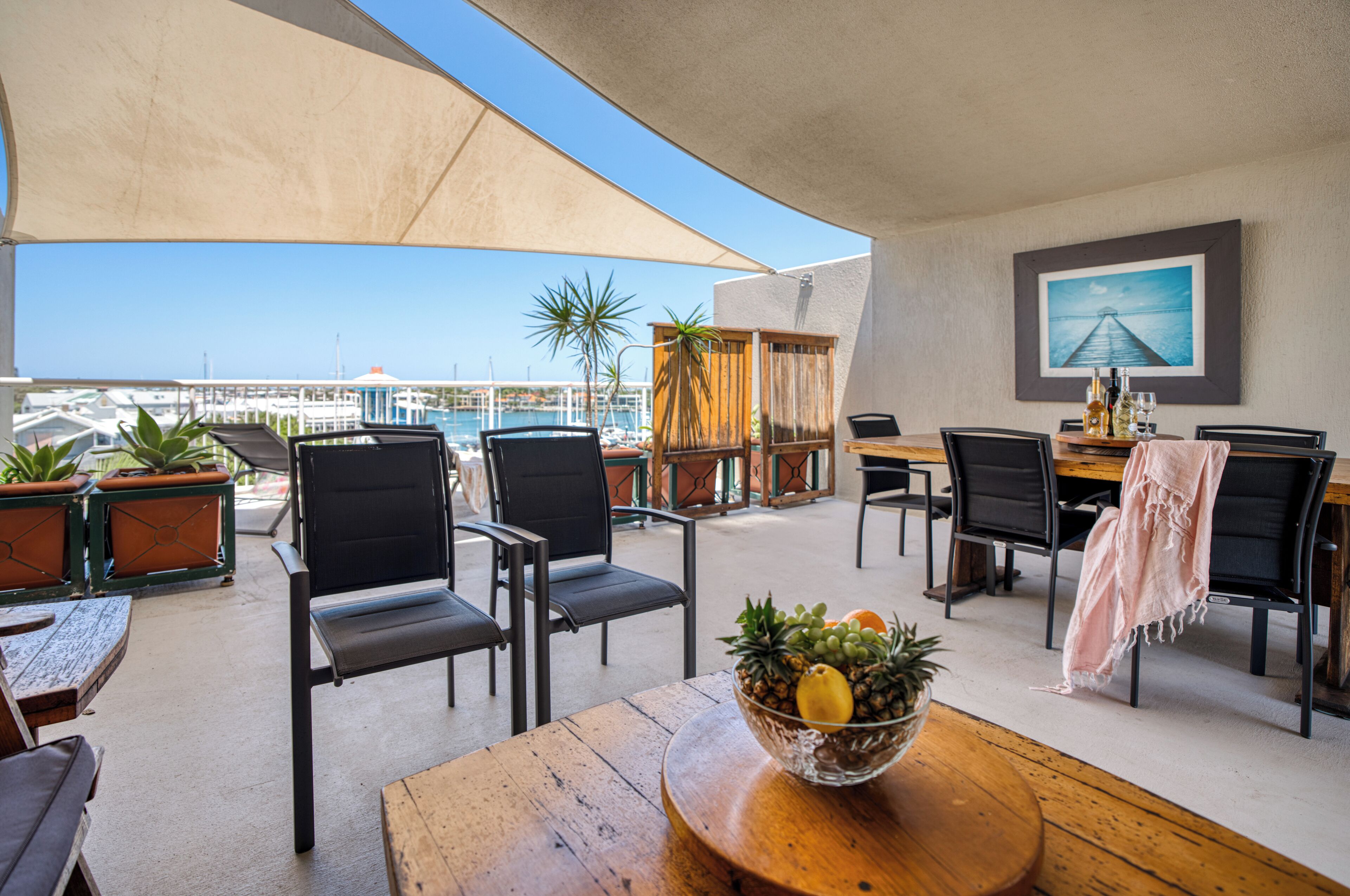 An outdoor patio with wooden tables and chairs, a large umbrella, and a view of the marina.