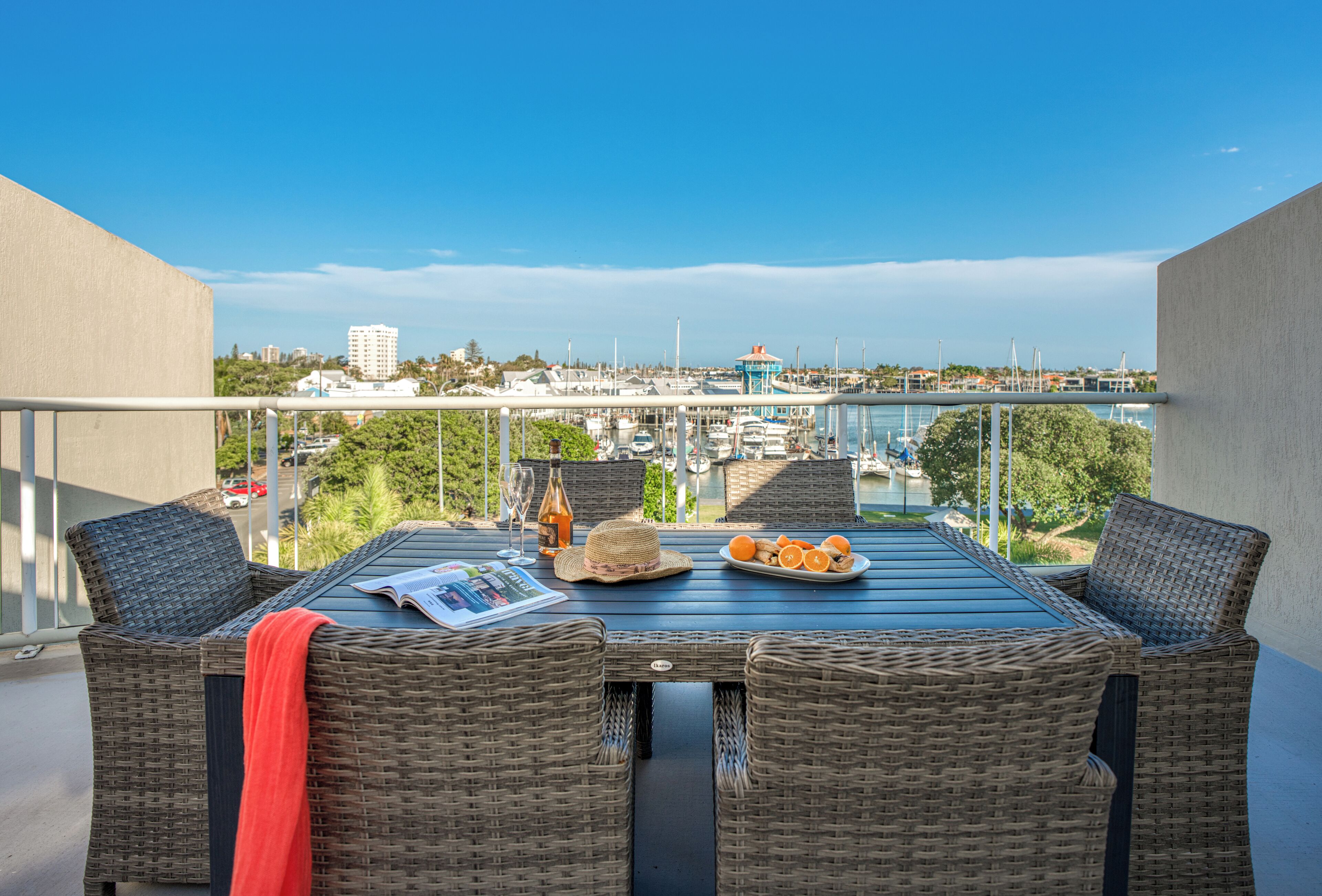 A balcony with wicker furniture, a table with a newspaper, a bottle, and a plate of oranges.